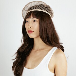 May include: A woman with long brown hair is wearing a white tank top and a beige mesh beret.