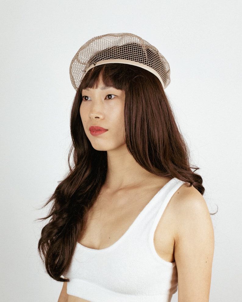 May include: A woman with long brown hair is wearing a white tank top and a beige mesh beret.