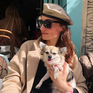 May include: A woman wearing a beige beret with a black mesh top and sunglasses is holding a small, white and brown chihuahua with a pink collar. The dog is sitting on the woman's lap and looking at the camera.