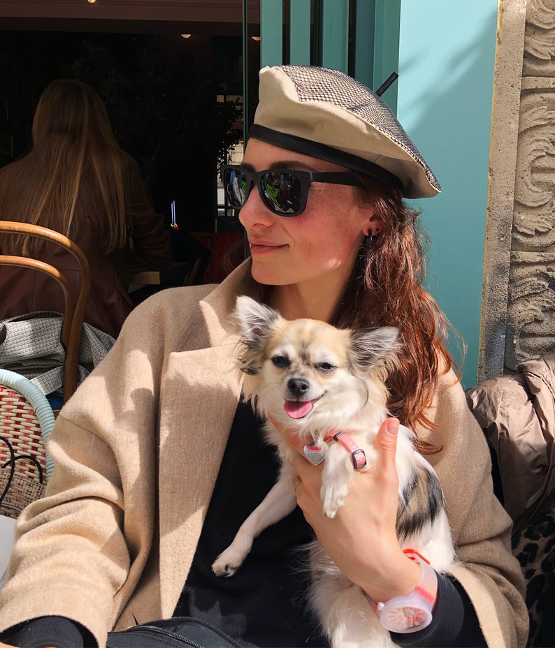 May include: A woman wearing a beige beret with a black mesh top and sunglasses is holding a small, white and brown chihuahua with a pink collar. The dog is sitting on the woman's lap and looking at the camera.