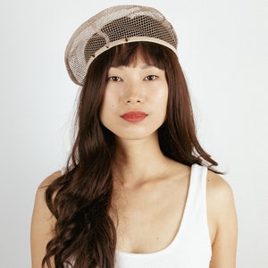 May include: A woman with long brown hair is wearing a beige mesh beret. She is wearing a white tank top.