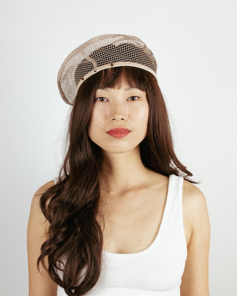 May include: A woman with long brown hair is wearing a beige mesh beret. She is wearing a white tank top.