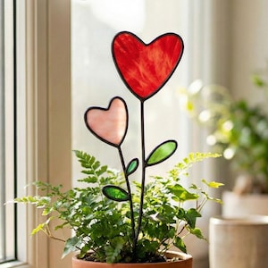 Puede incluir: Una planta en maceta con una flor decorativa de metal en forma de corazón. La flor tiene un corazón rojo y otro rosa, con hojas verdes. La planta está en una maceta de terracota, cerca de una ventana.