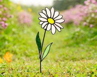 May include: A white daisy with a yellow center, made of stained glass, is attached to a black metal stem with green leaves. The daisy is standing in a bed of green grass.