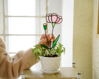 May include: A stained glass pink carnation flower with green leaves in a white ceramic pot with a green plant. The flower is being held by a hand.