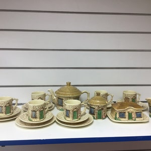 Puede incluir: Un juego de té vintage de temática de cabaña con una tetera, jarra de crema, azucarero y cuatro tazas y platillos. El juego está hecho de cerámica y presenta un diseño de cabaña con acentos marrones, blancos y verdes.