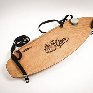 May include: A wooden surfboard with a black strap and a metal container. The board has a light brown finish with a black graphic of a mountain scene and the word "Elmo". The text "Corlette Surf" is also visible.