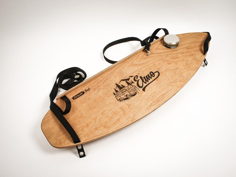 May include: A wooden surfboard with a black strap and a metal container. The board has a light brown finish with a black graphic of a mountain scene and the word "Elmo". The text "Corlette Surf" is also visible.