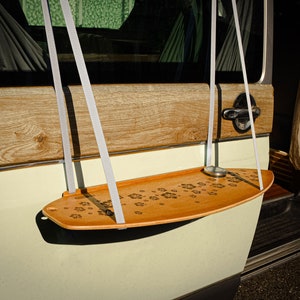 May include: A wooden swing seat with a paw print design hangs from white straps. The swing is attached to a vehicle's door. The seat is light brown and the vehicle is painted in a light green color.
