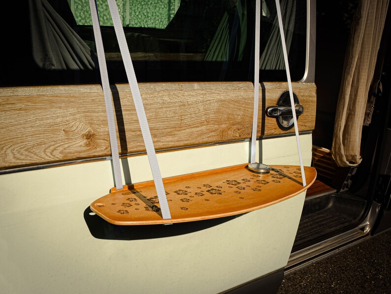 May include: A wooden swing seat with a paw print design hangs from white straps. The swing is attached to a vehicle's door. The seat is light brown and the vehicle is painted in a light green color.