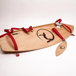 May include: A wooden surfboard with red straps and metal hardware. The board features a black graphic of a surfer inside a circle and the text "Curlsville Surf". A wooden tag with a surfer logo hangs from the board.