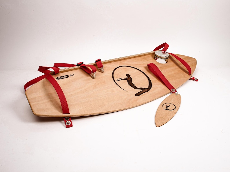 May include: A wooden surfboard with red straps and metal hardware. The board features a black graphic of a surfer inside a circle and the text "Curlsville Surf". A wooden tag with a surfer logo hangs from the board.