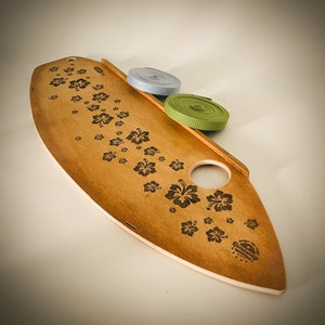 May include: A wooden surfboard-shaped serving board with a natural finish, adorned with etched hibiscus flower patterns. It features a round hole and a handle. Two rolls of ribbon, one light blue and one green, are placed on the board.