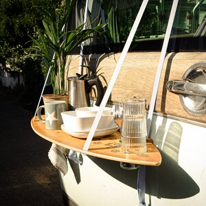 May include: A wooden serving tray with a surfboard shape is suspended from a vehicle's door. It holds a mug, bowls, plates, glasses, and silverware. A potted plant and a metal coffee pot are also on the tray. The door has a wooden panel and a chrome handle.