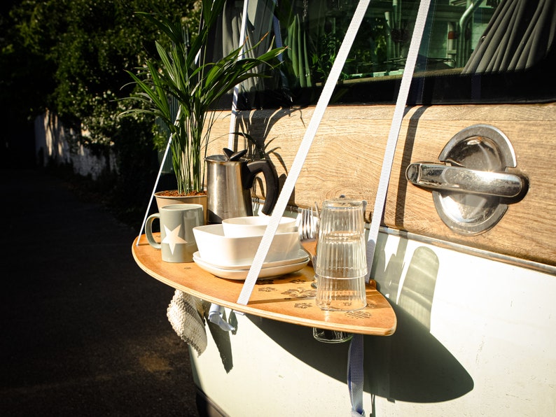 May include: A wooden serving tray with a surfboard shape is suspended from a vehicle's door. It holds a mug, bowls, plates, glasses, and silverware. A potted plant and a metal coffee pot are also on the tray. The door has a wooden panel and a chrome handle.