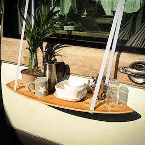 May include: A wooden surfboard-shaped shelf suspended by white straps, holding a potted plant, coffee pot, mug, bowls, cutlery, and a glass. The shelf is decorated with paw print designs and is attached to a vehicle.