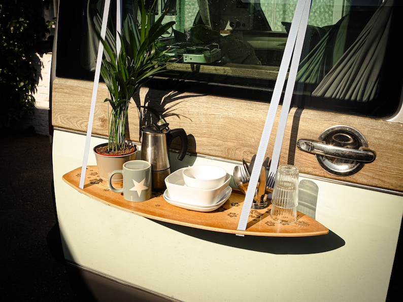 May include: A wooden surfboard-shaped shelf suspended by white straps, holding a potted plant, coffee pot, mug, bowls, cutlery, and a glass. The shelf is decorated with paw print designs and is attached to a vehicle.