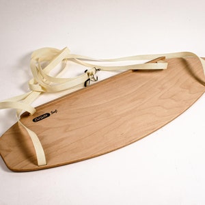 May include: A light-colored wooden skimboard with a cream-colored strap and the text "Carlsbad Surf". The board has an oval shape and is designed for skimming across the water's surface. The strap is attached to the board with metal hardware.