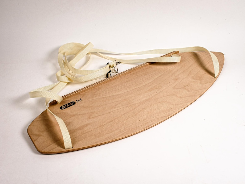 May include: A light-colored wooden skimboard with a cream-colored strap and the text "Carlsbad Surf". The board has an oval shape and is designed for skimming across the water's surface. The strap is attached to the board with metal hardware.