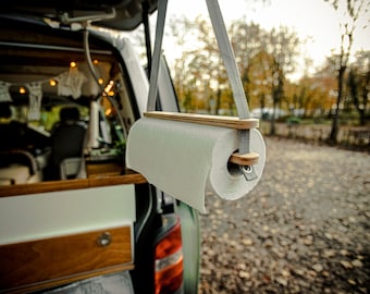 May include: A hanging paper towel holder made of light wood and gray straps. The holder is suspended from a vehicle's open back door, with a roll of white paper towels in place. The background shows a blurred outdoor scene.