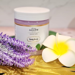 May include: A white jar of lavender vanilla body scrub with a gold label that says "Naturally Aglow Est. 2017" and "Body Scrub". The jar is sitting on a gold leaf with purple flowers.