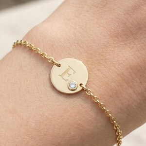 Könnte beinhalten: Goldfarbenes Armband mit einem kreisförmigen Anhänger, auf dem der Buchstabe "E" eingraviert ist. Unter dem Buchstaben ist ein kleiner, klarer Edelstein gesetzt. Das Armband hat eine feine Kette und wird mit einem Verschluss befestigt.
