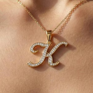 May include: A gold-coloured necklace featuring a pendant in the shape of the letter "K". The pendant is adorned with small, clear gemstones. The necklace is shown on a person's neck and chest.