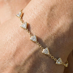 Puede incluir: Una delicada pulsera de oro con una serie de piedras preciosas triangulares y transparentes. La cadena de la pulsera es de color oro cálido, y las piedras preciosas están engastadas en biseles individuales de oro. El diseño es sencillo y elegante, adecuado para el uso diario o ocasiones especiales.