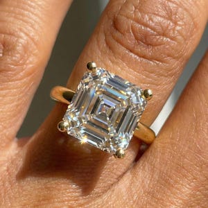 May include: A gold ring featuring a large, square-cut diamond. The diamond has a complex, multi-faceted design that reflects light. The ring has a simple, elegant band with small gold accents at each corner of the diamond.