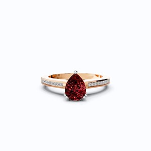 May include: A gold ring with a pear-shaped, deep red gemstone as the focal point. The band is accented with small, clear gemstones. The ring is set against a white background, highlighting its elegant design.