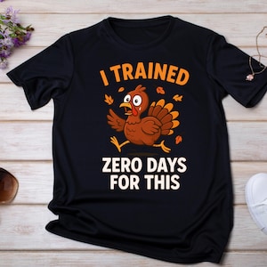 May include: Black t-shirt with a cartoon turkey graphic. The turkey is brown with orange accents and is running. The text on the shirt reads "I TRAINED" above the turkey and "ZERO DAYS FOR THIS" below.