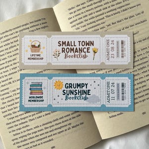 May include: Two colorful bookmarks with a ticket design. The top bookmark is beige with a floral design and the text "Small Town Romance Bookclub". The bottom bookmark is blue with a yellow sun and the text "Grumpy Sunshine Bookclub".