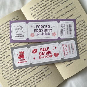 May include: Two colorful bookmarks with a ticket design. The top bookmark is purple and says "Forced Proximity Bookclub" with a pink flower on each side. The bottom bookmark is gray and says "Fake Dating Bookclub" with a red heart on each side. Both bookmarks have a red star on each side.