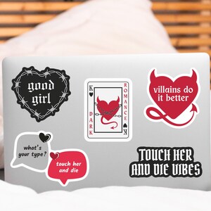 May include: A laptop covered in stickers with various designs and text. Stickers include a heart in barbed wire with "good girl", a playing card with a chained heart, and a red heart with "villains do it better". Other stickers say "what's your type?" and "touch her and die".