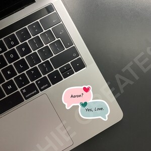 May include: A laptop computer with a pink and blue speech bubble sticker with the text "Aaron?" and "Yes, Love."