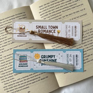 May include: Three different bookmark designs with tassels. The bookmarks feature illustrations of a book stack, a sun, and a flower. The text on the bookmarks reads "Lifetime Membership", "Worldwide Membership", "Small Town Romance", "Grumpy Sunshine", "Admit One 23 08 24", and "Admit One 1 07 24".