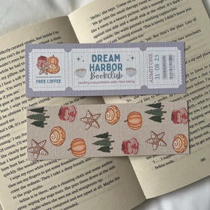 May include: A bookmark with a grey and white design featuring a book club logo with the text "Dream Harbor Bookclub" and the words "reading was probably safer than hiking". The bookmark is decorated with a pattern of autumn-themed illustrations including pumpkins, cinnamon rolls, and pine trees.
