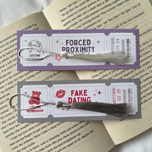May include: Two purple and white bookmarks with silver tassels. The bookmarks feature different designs and text. The first bookmark says "Forced Proximity" and the second bookmark says "Fake Dating".