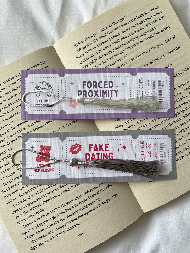 May include: Two purple and white bookmarks with silver tassels. The bookmarks feature different designs and text. The first bookmark says "Forced Proximity" and the second bookmark says "Fake Dating".