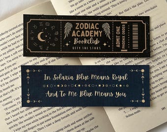 May include: A black bookmark with gold lettering and a celestial design. The bookmark features the text "Zodiac Academy Bookclub" and "Defy the Stars". The bookmark also includes the text "In Solaria Blue Means Royal" and "And To Me Blue Means You".