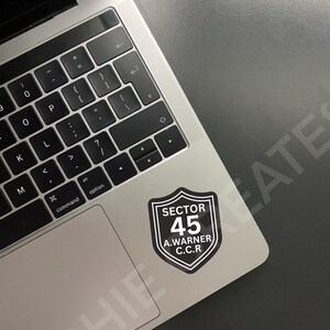 May include: A black and white sticker with a shield shape and the text "SECTOR 45 A. WARNER C.C.R." on a silver laptop.