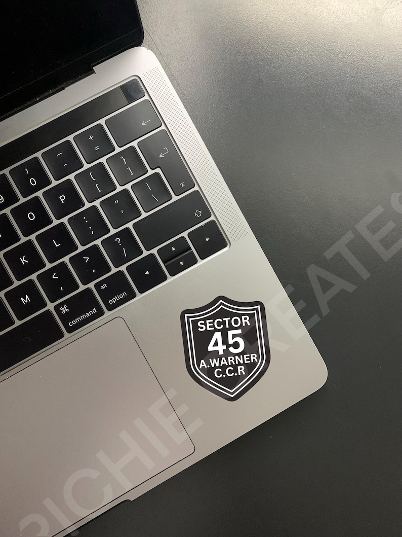 May include: A black and white sticker with a shield shape and the text "SECTOR 45 A. WARNER C.C.R." on a silver laptop.