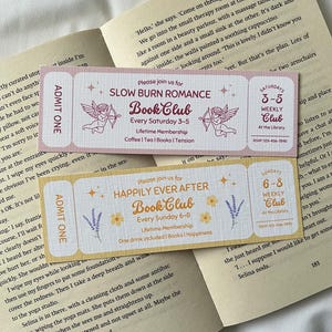 May include: Two pink and yellow bookmarks with a whimsical design. The bookmarks feature illustrations of cherubs with arrows and text that reads "Slow Burn Romance Book Club" and "Happily Ever After Book Club".