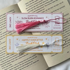 May include: Two pink and white bookmark tickets with tassels. The pink bookmark has the text "Please join us for Slow Burn Romance Book Club" and the white bookmark has the text "Please join us for Happily Ever After Book Club".