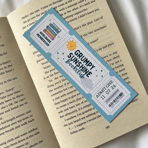 May include: A blue and white bookmark with a sun and the text "Grumpy Sunshine Bookclub" on it. The bookmark also has a ticket-style design with the text "Admit One 11 07 24" and a barcode.