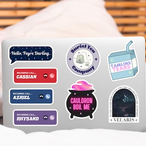 May include: A laptop computer with several colorful stickers featuring text and illustrations. The stickers include a cauldron with the text "Cauldron Boil Me", a box with the text "Tamlin's Tears", a speech bubble with the text "Hello, Feyre Darling", and a sticker with the text "Suriel Tea Company".