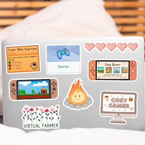 May include: A laptop computer with several stickers on it. The stickers are of a video game controller, a campfire, a video game console, a coffee cup, and a plant. The text on the stickers reads "Gamer", "Cozy Gamer", "Virtual Farmer", and "Cozy Gamer".