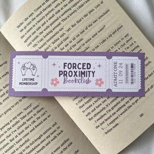May include: Purple and white bookmark with the text "Forced Proximity Bookclub" and a hand holding a heart. The bookmark also has a barcode and the text "Admit One 11 09 24".