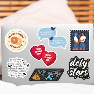 May include: A silver laptop with various stickers, including a heart with the text "There is only him", a heart with the text "There is only her", a sticker with the text "Buttery Bagels Geraldine's", a sticker with the text "In solaria blue means royal", a sticker with the text "And to me, blue means you", a sticker with the text "Defy the stars", a sticker with the text "wolfman bitey c bitchy flame eyes dragzilla phoen dream proffesor shame fish fury batty betty", and a sticker with four triangles, each with a different element: fire, earth, water, and air.