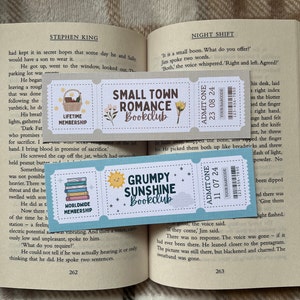 May include: Two colorful bookmarks with a book club theme. The top bookmark is light blue with a white and brown illustration of a small town and the text "Small Town Romance Bookclub". The bottom bookmark is light blue with a yellow sun and the text "Grumpy Sunshine Bookclub".
